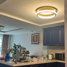 Niebieska zabudowa kuchenna z marmurowym blatem, ekspres do kawy, okrągła lampa sufitowa LED z złotymi akcentami, kartonowe pudło na podłodze.