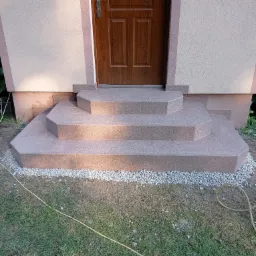 Wykończone, kafelkowane schody wejściowe do domu z brązowymi drzwiami, otoczone białym żwirem i zielonym trawnikiem.