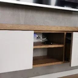 Nowoczesna szafka RTV z białymi, lakierowanymi frontami i drewnianym blatem, z widocznymi gniazdkami elektrycznymi w otwartej przestrzeni, na tle szarej ściany i drewnianych paneli.