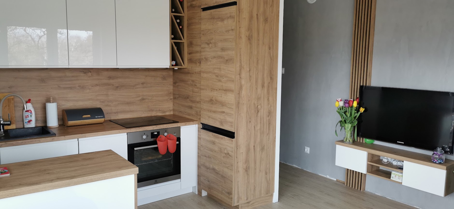 Nowoczesna kuchnia z białymi frontami szafek i drewnianymi blatami, z akcentem w postaci szafki na wino i dekoracyjnych listew na ścianie, widok na salon z telewizorem i tulipanami.