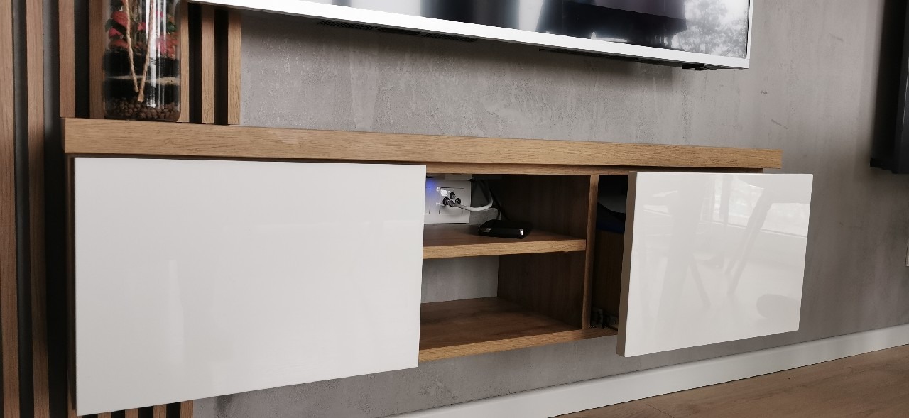 Nowoczesna szafka RTV z białymi, lakierowanymi frontami i drewnianym blatem, z widocznymi gniazdkami elektrycznymi w otwartej przestrzeni, na tle szarej ściany i drewnianych paneli.