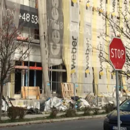 Budynek w trakcie ocieplania, z widocznym rusztowaniem, materiałami izolacyjnymi i banerami reklamowymi firm Weber i Adatex.pl, na pierwszym planie znak STOP i zaparkowany samochód oraz bus marki...