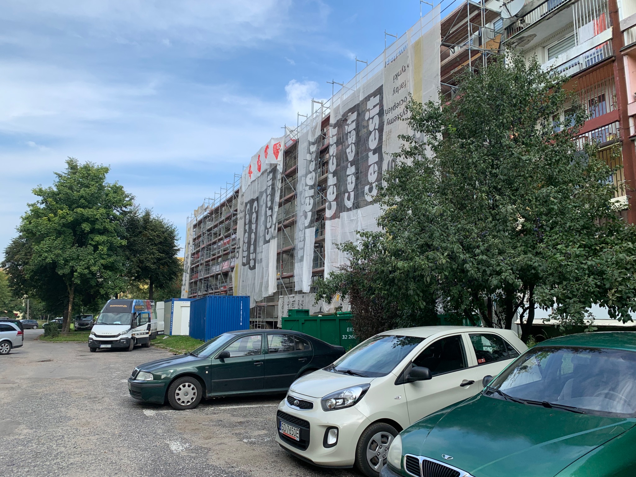 Budynek mieszkalny w trakcie ocieplania, osłonięty rusztowaniami i siatką z nadrukowanym logo producenta materiałów budowlanych, widok z parkingu z zaparkowanymi samochodami.
