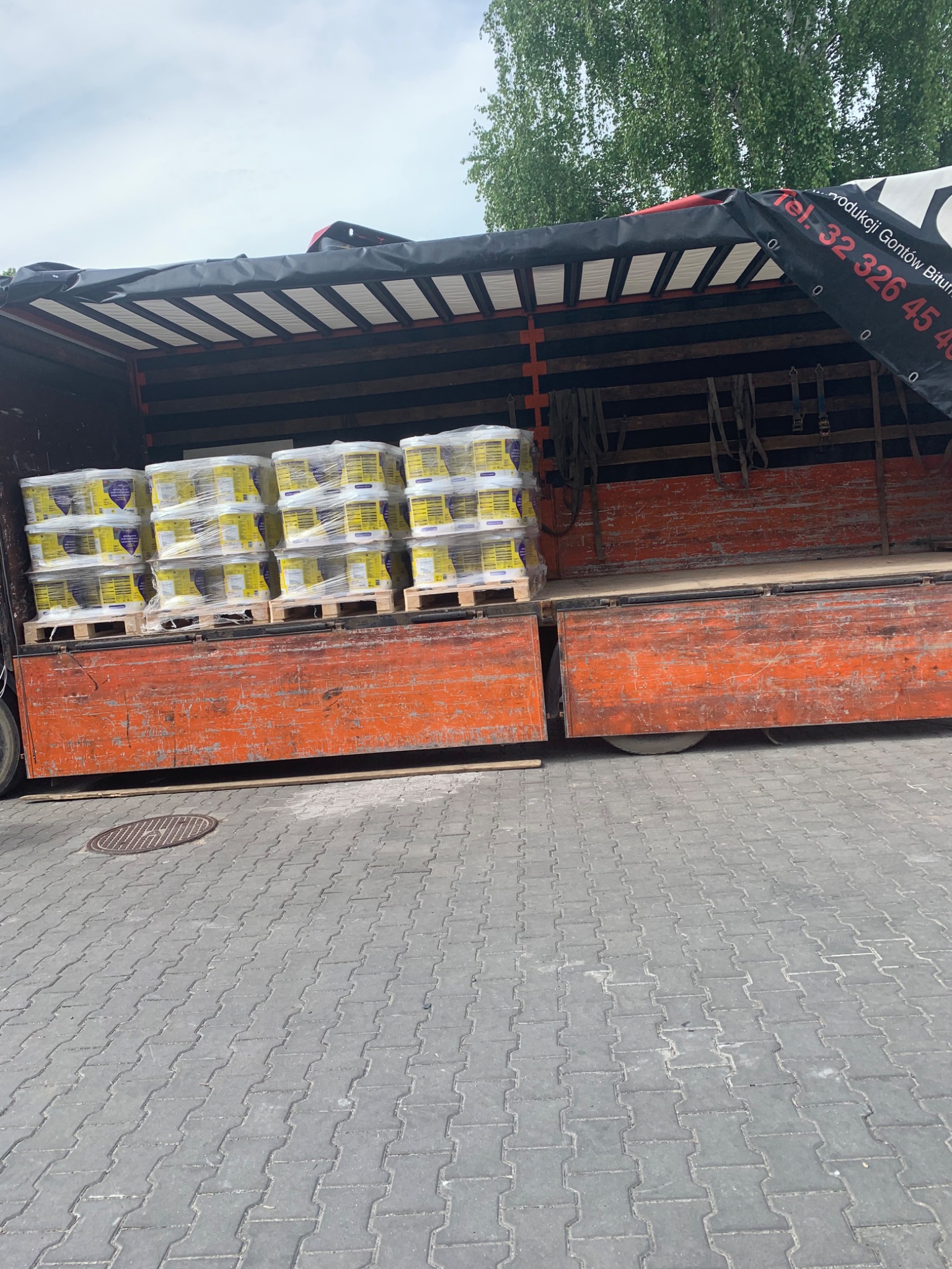 Widok z boku na otwartą naczepę ciężarówki z załadowanymi paletami z pojemnikami z materiałem budowlanym, zabezpieczonymi folią stretch, na tle szarej kostki brukowej.