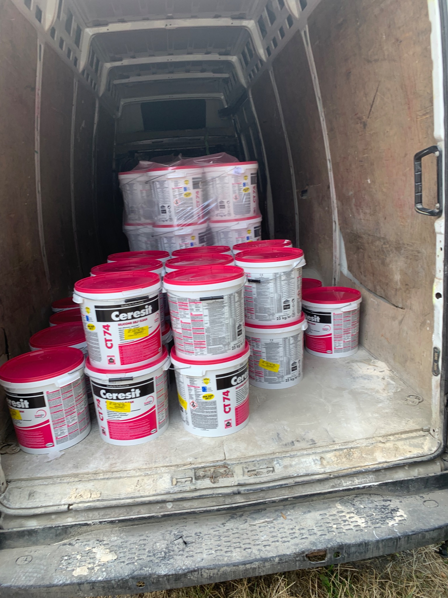 Wnętrze brudnego furgonetki wypełnione stosem wiader z zaprawą klejową marki Ceresit CT74, część wiader zapakowana w folię, widoczne ślady zaprawy na podłodze i ścianach pojazdu.