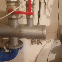 Szara, metalowa instalacja z czerwonymi zaworami i manometrami w piwnicy, z widocznymi rurami i przyłączami.