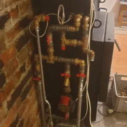 Kotłownia z piecem na paliwo stałe, widoczne rury miedziane i stalowe z zaworami, instalacja grzewcza przy ceglanej ścianie.