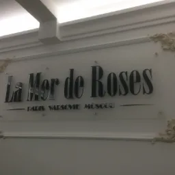Elegancki szyld z nazwą 'La Mer de Roses' w czcionce szeryfowej, umieszczony na białej ścianie z dekoracyjnymi narożnikami i podświetleniem sufitowym.