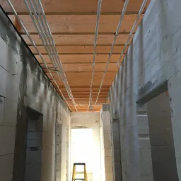 Wnętrze surowego domu w trakcie budowy, widoczne ściany z betonu komórkowego, drewniany strop z instalacją elektryczną w białych rurach, żółta drabina w przejściu.