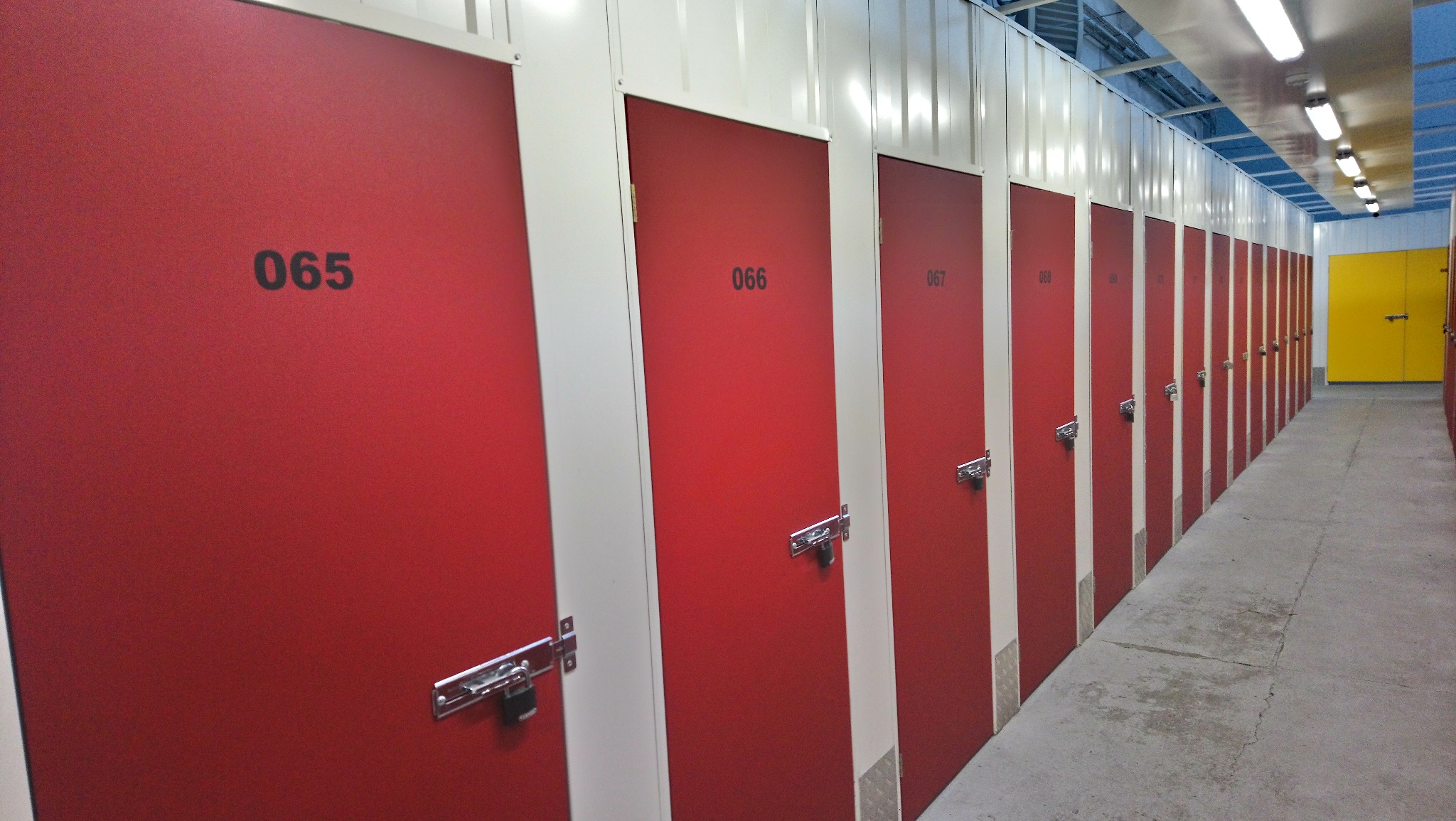 Rząd czerwonych, ponumerowanych boksów zamykanych na kłódkę w magazynie samoobsługowym, widok z perspektywy korytarza, z żółtym boksem w tle.