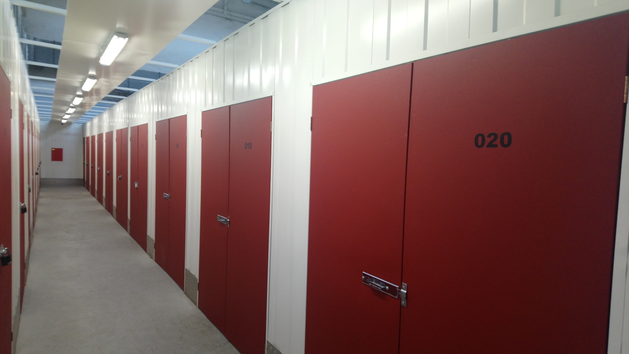 Korytarz magazynu samoobsługowego z rzędem czerwonych boksów oznaczonych numerami, oświetlony rzędami lamp na suficie.