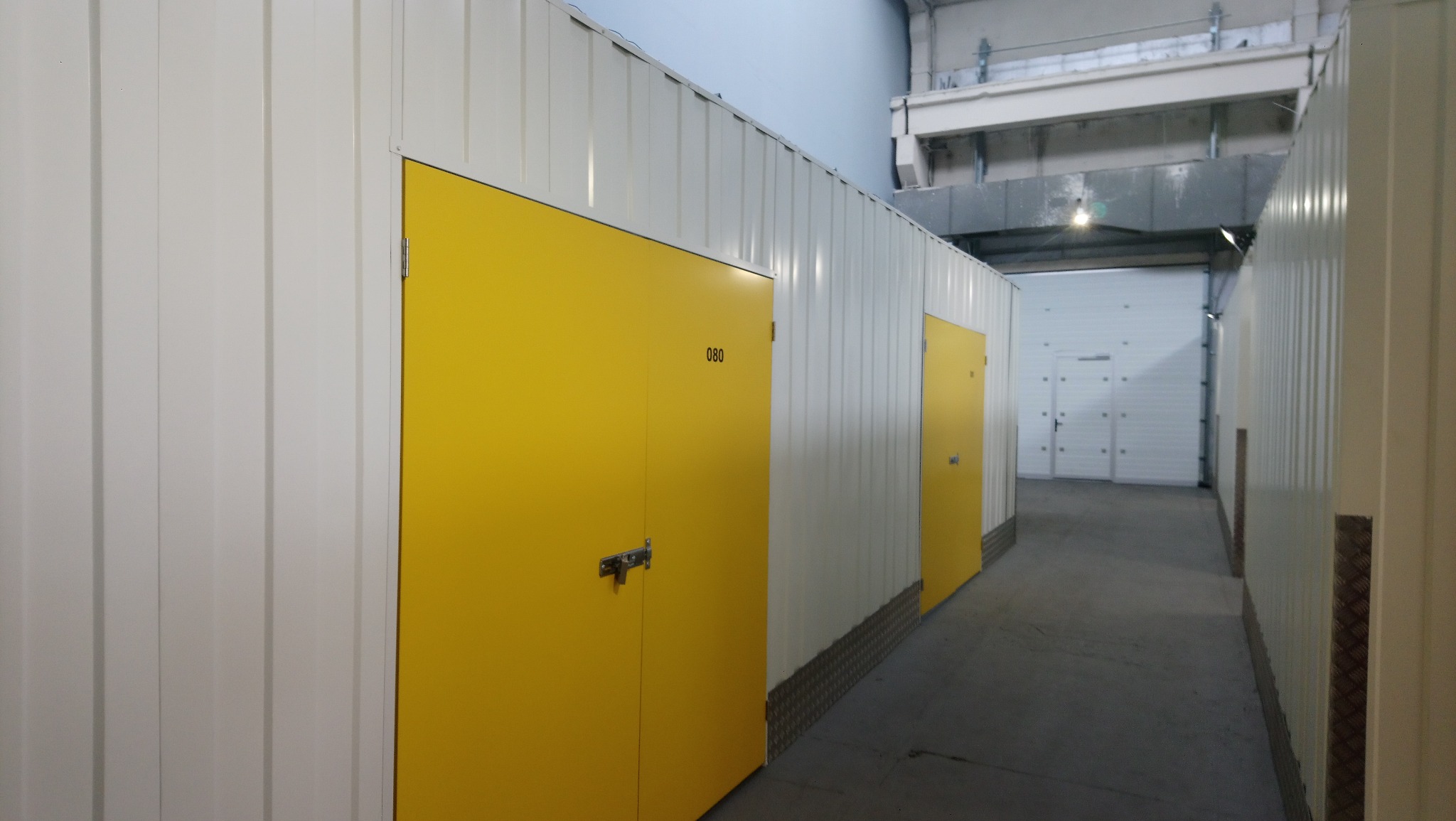 Rząd boksów w magazynie samoobsługowym, z żółtymi drzwiami oznaczonymi numerem 080, widok z perspektywy korytarza wewnątrz magazynu.