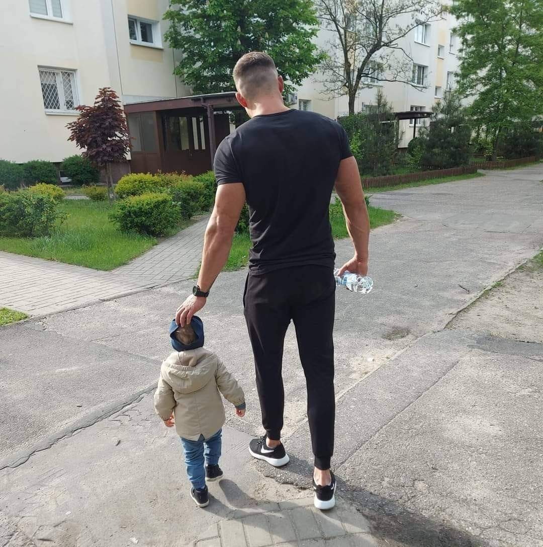 Mężczyzna w czarnym ubraniu trzymający za głowę małego chłopca na tle bloku mieszkalnego i zieleni.