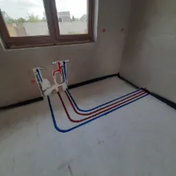 Wewnętrzny kąt pomieszczenia w trakcie instalacji hydraulicznej: widoczne rury ciepłej i zimnej wody w kolorach czerwonym i niebieskim, prowadzone po podłodze w kierunku ściany z przygotowanymi...