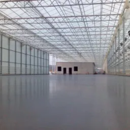 Błyszcząca, szara posadzka przemysłowa wewnątrz szklarni z metalową konstrukcją i białymi ścianami bocznymi. W tle widoczny budynek z otworami okiennymi.