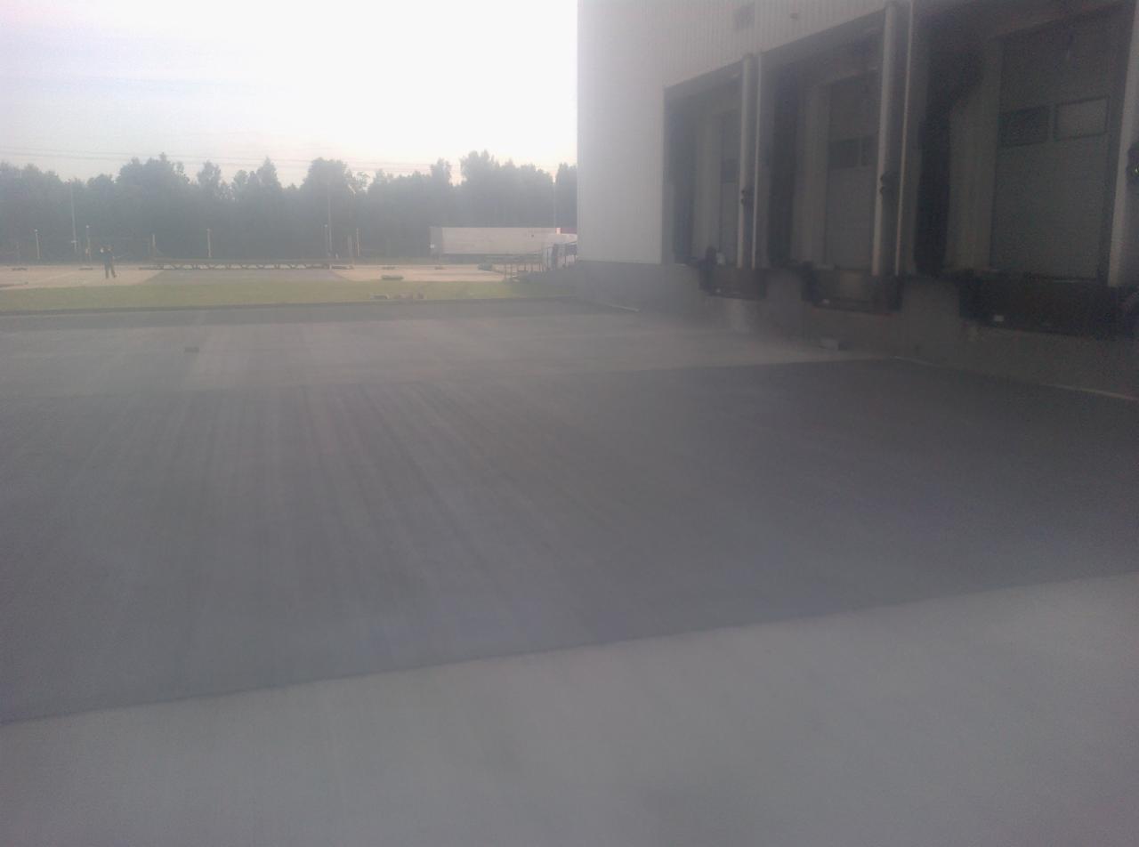 Szara, gładka posadzka przemysłowa przed budynkiem magazynowym z rampami załadunkowymi w tle. Widoczny fragment zieleni i drzewa.