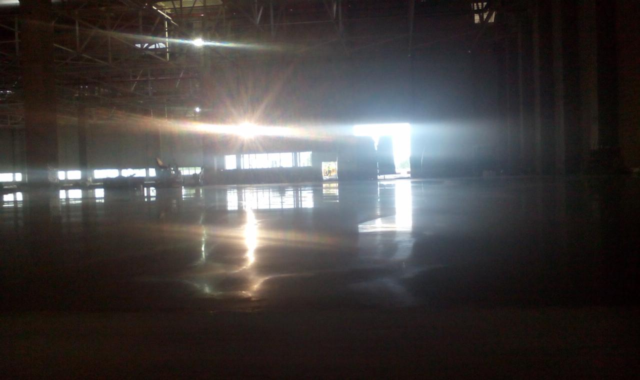 Błyszcząca posadzka przemysłowa w hali z widocznymi odbiciami światła słonecznego wpadającego przez okna i drzwi. Widoczne elementy konstrukcji dachu.