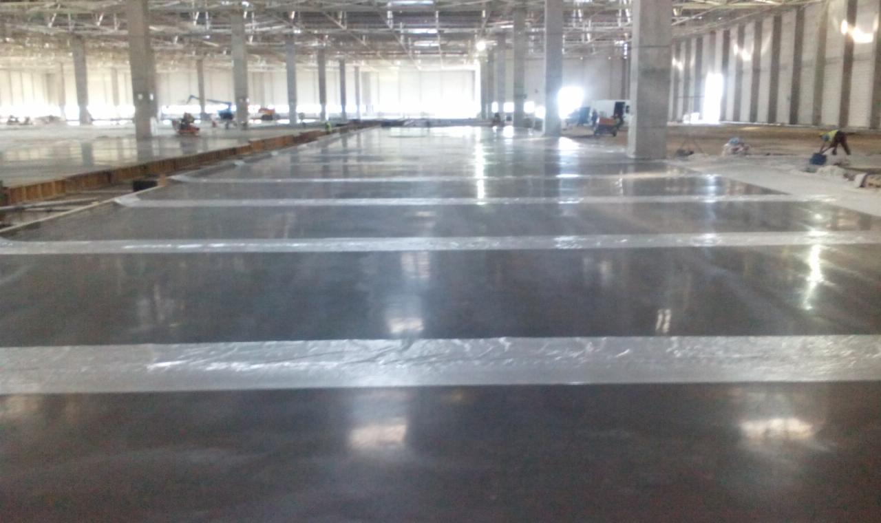 Błyszcząca, ciemna posadzka przemysłowa w hali produkcyjnej z widocznymi pasami zabezpieczającymi i pracownikami w oddali. Widok z niskiego kąta.