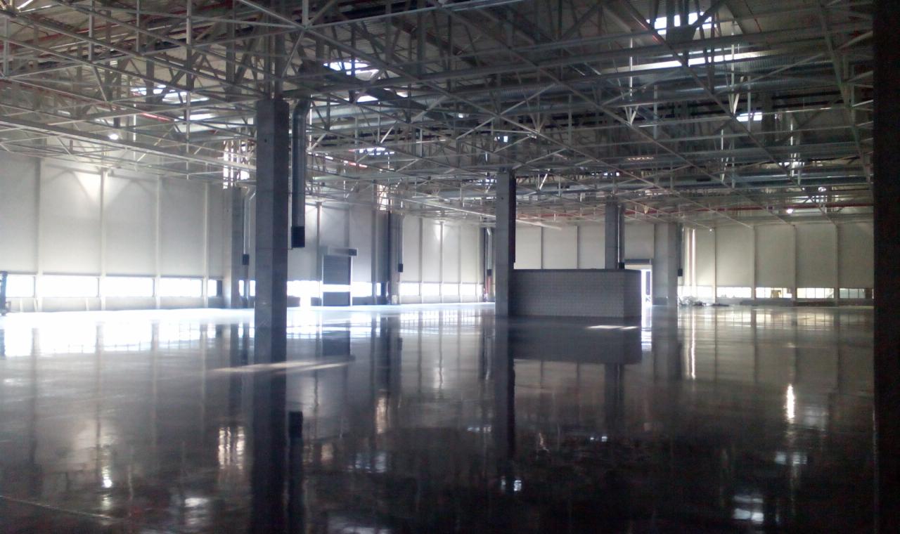 Błyszcząca, ciemna posadzka przemysłowa w przestronnej hali z metalową konstrukcją sufitu i kolumnami. Światło wpada przez okna, tworząc refleksje na podłodze.