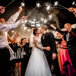 Pocałunek nowożeńców otoczonych gośćmi z zimnymi ogniami podczas wesela; ciemne tło, ciepłe światło.