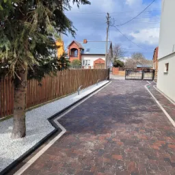 Ułożona z brązowej kostki brukowej droga dojazdowa z białym kamieniem ozdobnym wzdłuż ogrodzenia i drzewa iglastego, brama wjazdowa w tle.