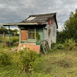 Zrujnowana, mała budowla z blachy falistej i elementami drewnianymi, z widocznymi śladami korozji i odpadającej farby, otoczona zarośniętym terenem i porozrzucanymi elementami budowlanymi.