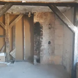 Pokój w trakcie remontu: odsłonięta cegła komina z czarnymi śladami sadzy, drewniane belki stropowe, częściowo otynkowane ściany, podłoga po wylewce.