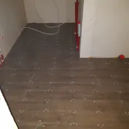 Ułożone panele podłogowe imitujące drewno, z białymi krzyżykami dystansowymi, widoczne rury instalacyjne na ścianie i podłodze, fragment surowego muru w rogu.