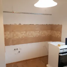 Fragment ściany w kuchni w trakcie kładzenia płytek, widoczne niedociągnięcia i pozostałości zaprawy, na podłodze resztki materiałów, w tle biała zabudowa kuchenna z piekarnikiem.