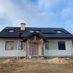 Montaż paneli słonecznych na dachu domu jednorodzinnego; pracownik na drabinie przy kominie, elewacja w trakcie wykończenia, okna zabezpieczone folią.
