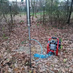 Próbnik geologiczny wbijany w ziemię na tle opadłych liści, obok czerwony kompresor i tablet z widocznym odbiciem nieba.