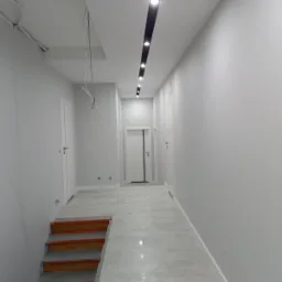 Szary korytarz z drewnianymi schodami zabezpieczonymi taśmą malarską, szare płytki na podłodze, białe ściany i drzwi, oraz czarna listwa oświetleniowa na suficie.