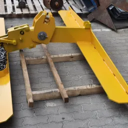 Żółty, metalowy uchwyt do podnoszenia z łańcuchem, leżący na drewnianej palecie na kostce brukowej.