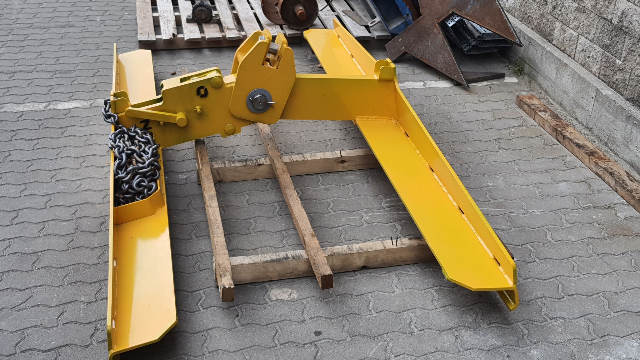 Żółty, metalowy uchwyt do podnoszenia z łańcuchem, leżący na drewnianej palecie na kostce brukowej.