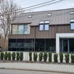 Nowoczesny budynek kliniki dentystycznej w Krakowie z brązową elewacją, rzędem małych drzewek przed wejściem i zaparkowanym niebieskim samochodem w tle.