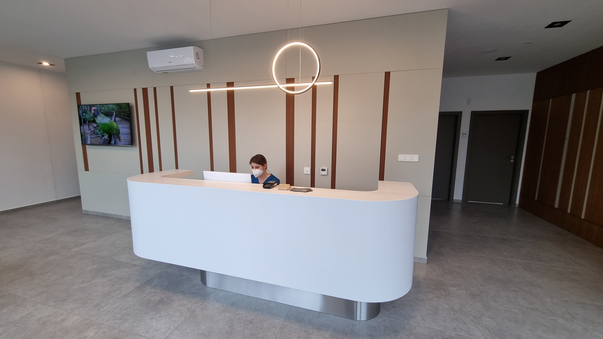 Nowoczesna recepcja w klinice medycznej z pracownikiem w maseczce za białym, zakrzywionym biurkiem. W tle widoczny telewizor z motywem roślinnym i minimalistyczne oświetlenie.