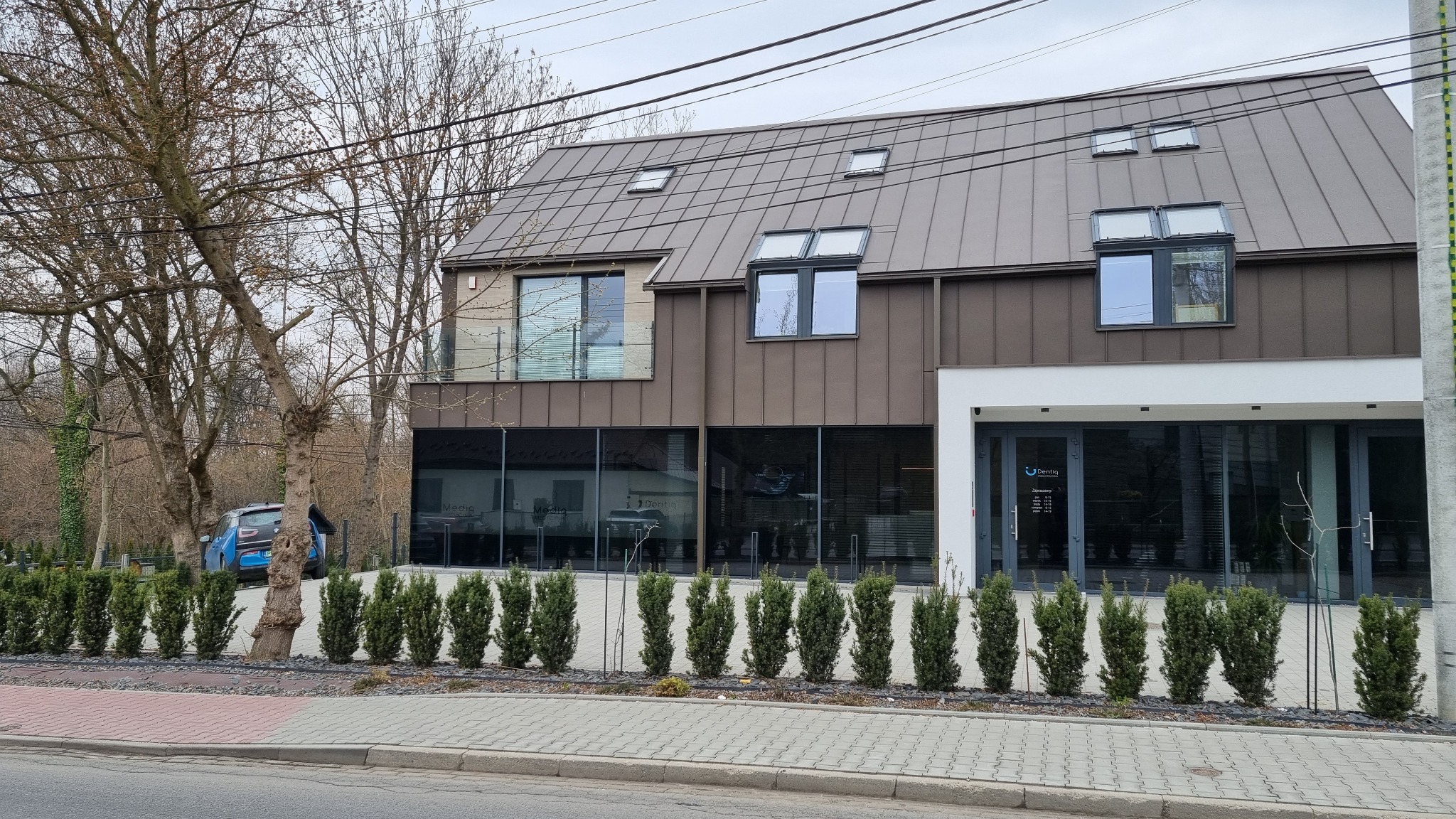 Nowoczesny budynek kliniki dentystycznej w Krakowie z brązową elewacją, rzędem małych drzewek przed wejściem i zaparkowanym niebieskim samochodem w tle.