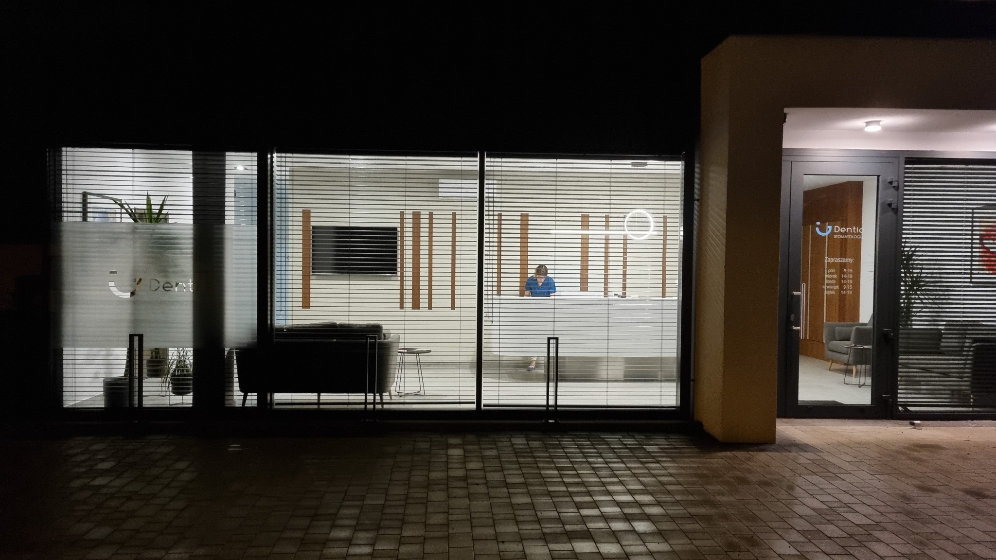 Widok z zewnątrz na recepcję kliniki dentystycznej nocą, z widoczną pracownicą za biurkiem, logo na szybie i godzinami otwarcia na drzwiach wejściowych.