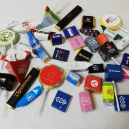 Różnorodność słodyczy i lizaków z nadrukami logo różnych firm, ułożone na jasnym tle, stanowiące gadżety reklamowe.