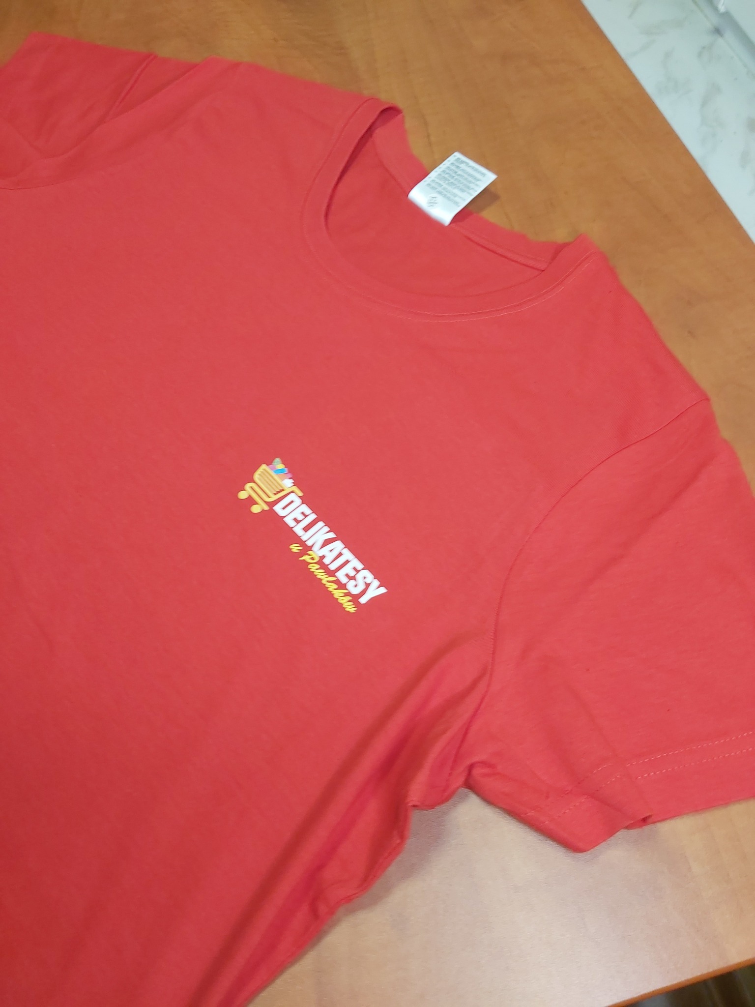 Czerwona koszulka z nadrukowanym logo 'Delikatesy u Pawlaków' w kolorach żółtym i białym, leżąca na drewnianym stole.