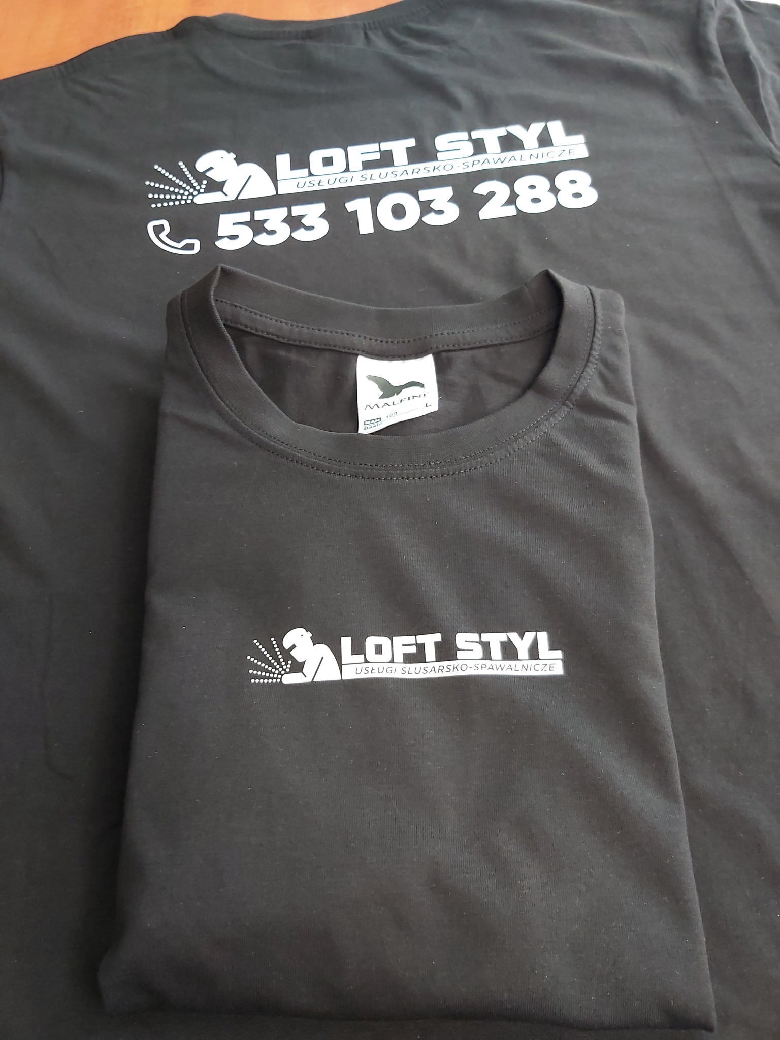 Czarne koszulki z nadrukowanym logo firmy 'Loft Styl' oferującej usługi ślusarsko-spawalnicze oraz numerem telefonu, ułożone na ciemnym tle.