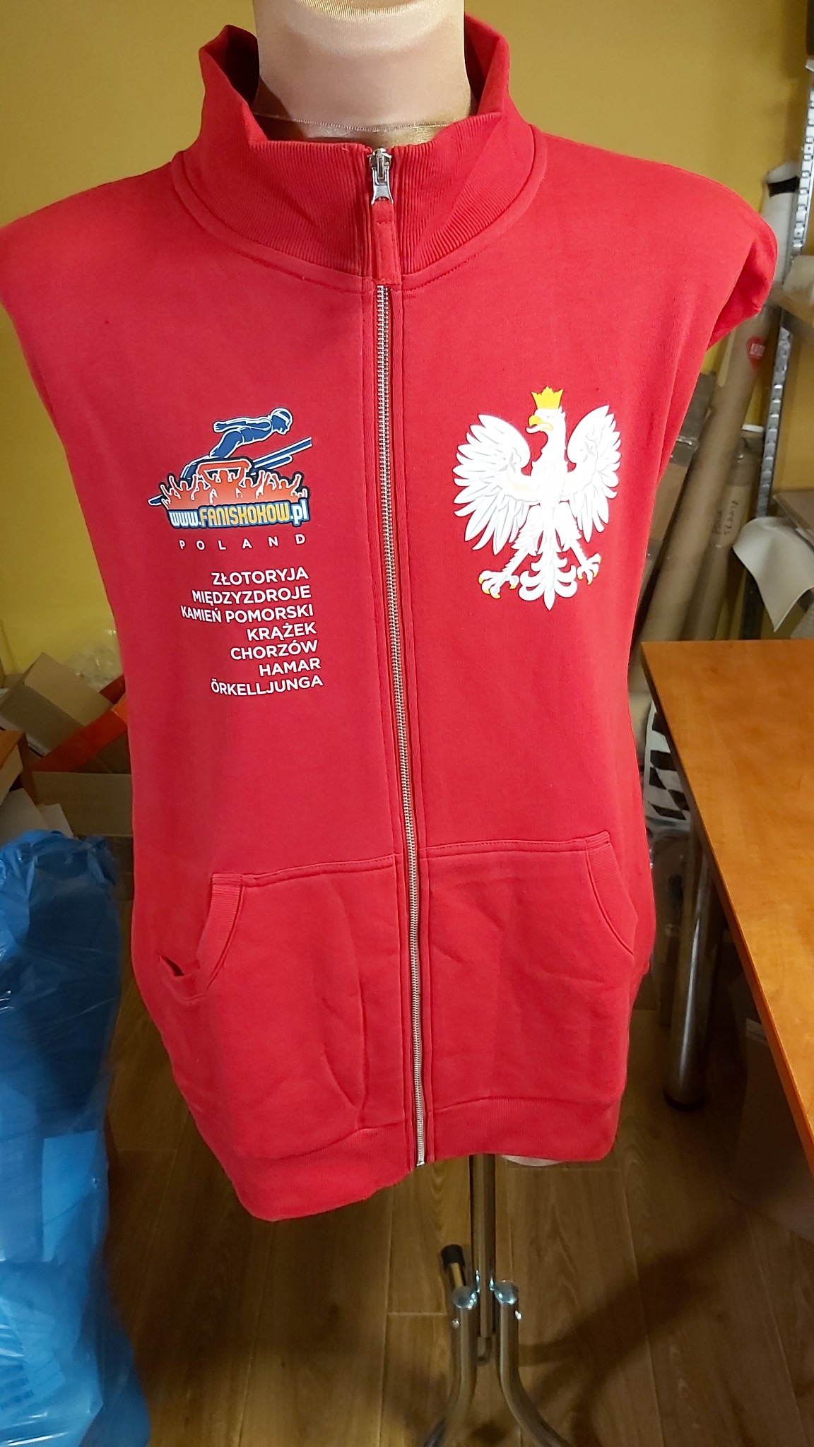 Czerwona bluza bez rękawów z zamkiem błyskawicznym, nadrukiem logotypu 'www.fanishow.pl' i godłem Polski na piersi, umieszczona na manekinie.