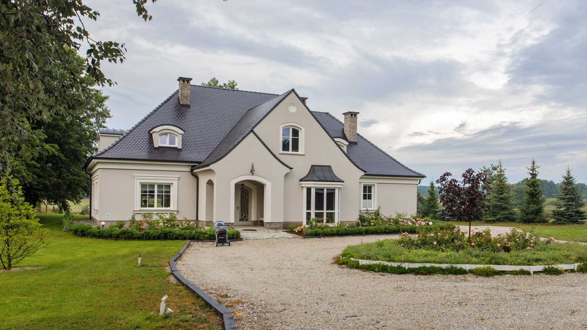 Elegancki dom jednorodzinny z ciemną dachówką, kremową elewacją i starannie zaprojektowanym ogrodem z krzewami róż, widok z perspektywy podjazdu z żwiru.