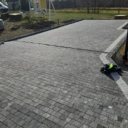 Ułożona, szara kostka brukowa na posesji z widoczną kratką odpływową, paletami kostki i domkiem dla dzieci w tle.