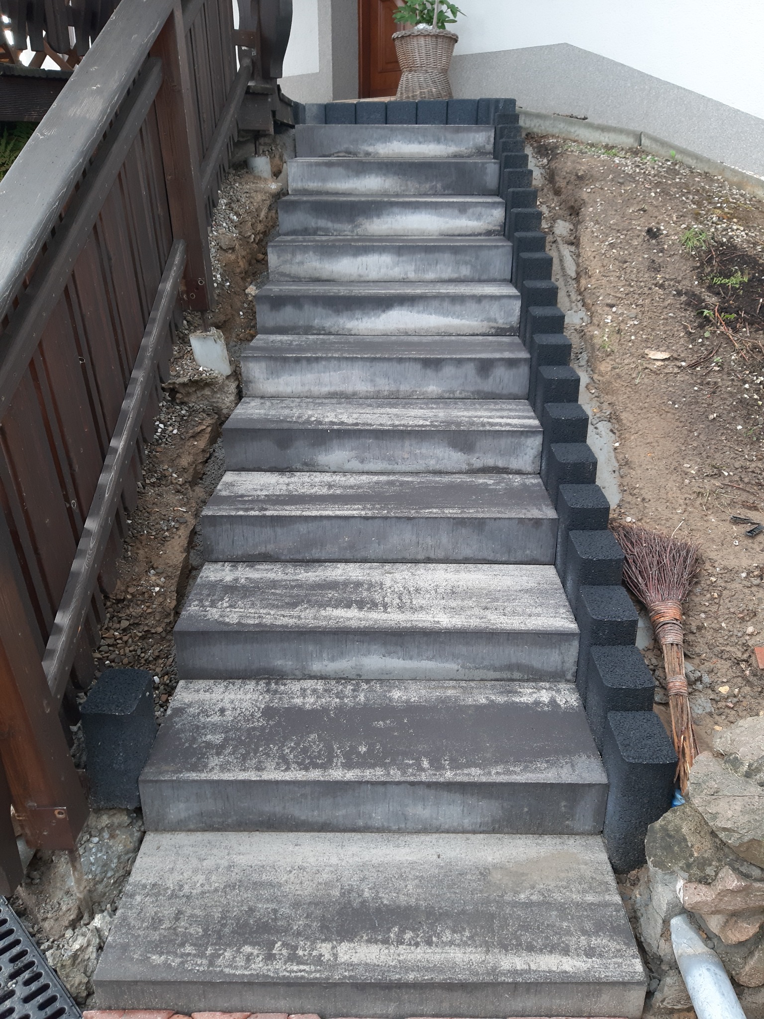 Schody zewnętrzne z szarych, betonowych płyt, obłożone czarną kostką po prawej stronie, prowadzące do wejścia domu z wiklinowym koszem na górnym stopniu.