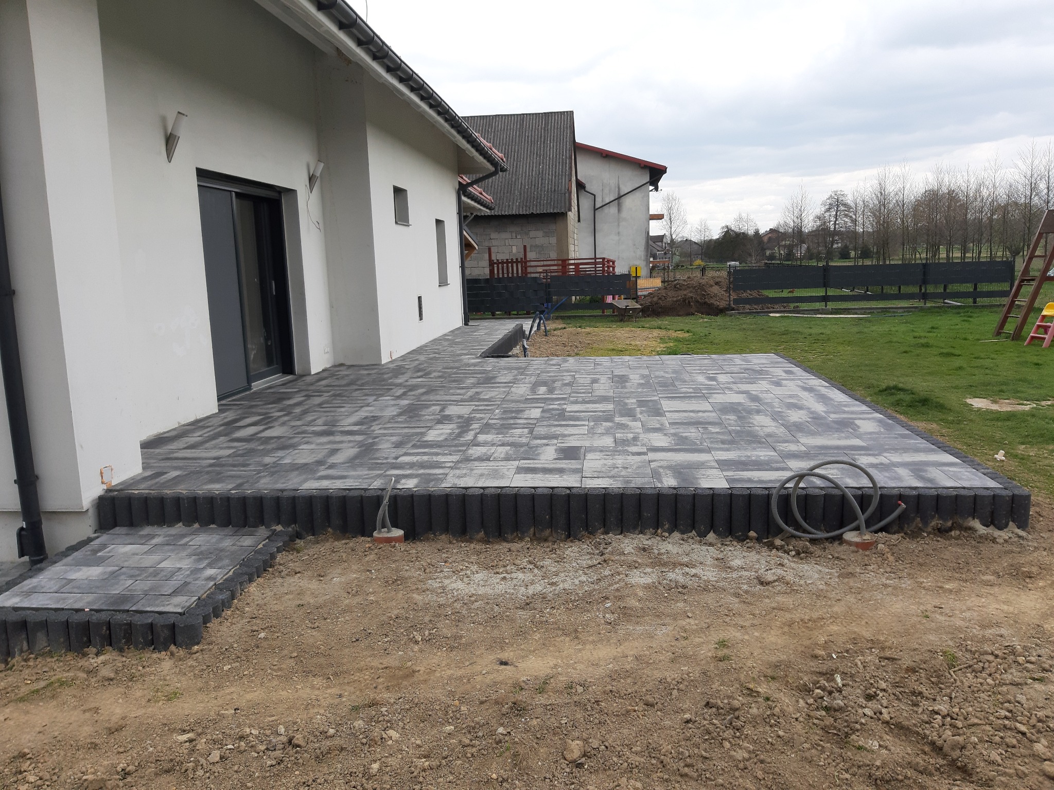 Nowoczesny taras z szarej kostki brukowej ułożony przy białym domu z przesuwnymi drzwiami, widoczne obrzeża i przygotowany teren.