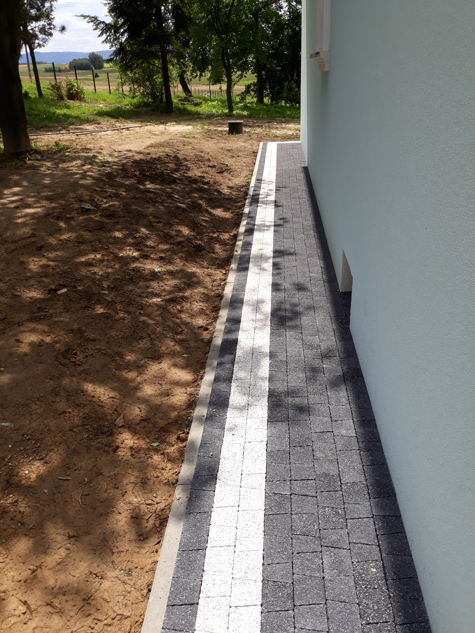 Świeżo ułożony chodnik z kostki brukowej w kolorze szaro-białym, biegnący wzdłuż ściany budynku z widocznym fragmentem trawnika i drzew w tle.