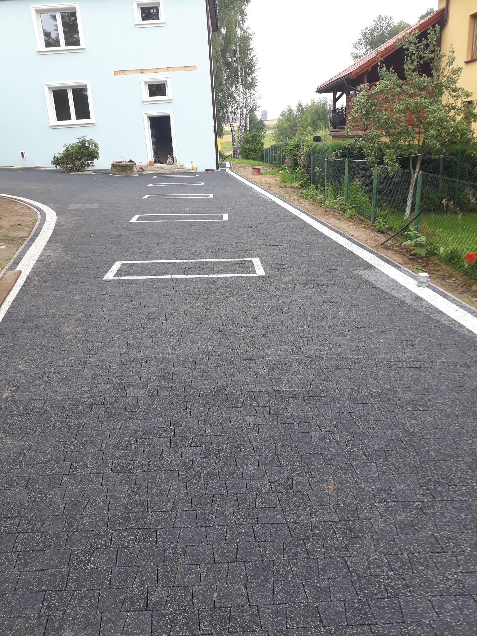 Czarna kostka brukowa ułożona na podjeździe z białymi obramowaniami i prostokątnymi wstawkami, widok na dom z jasnoniebieską elewacją i zielony ogród w tle.