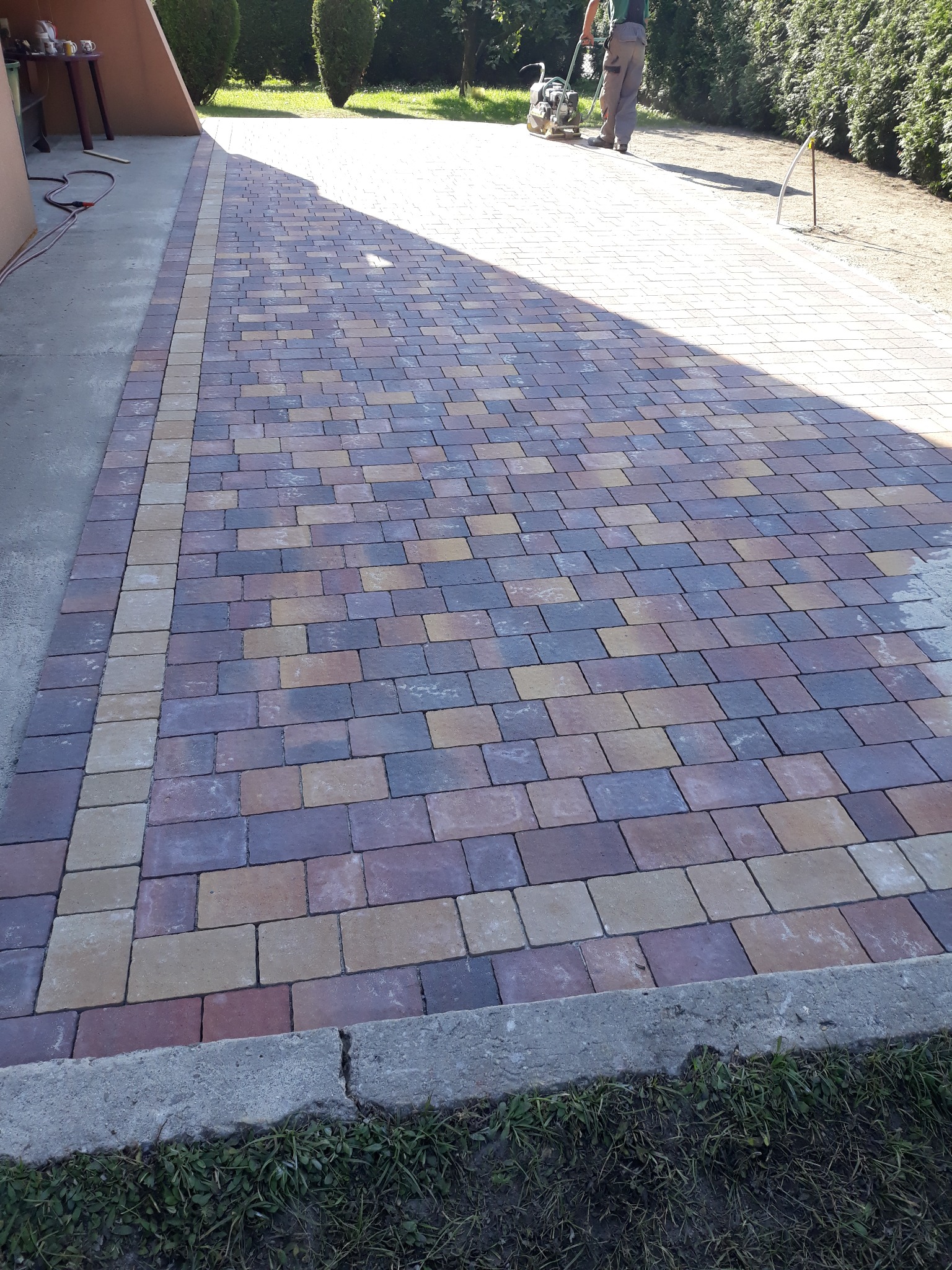 Nowo ułożona, wielobarwna kostka brukowa tworząca podjazd, z widocznym pracownikiem zagęszczającym powierzchnię przy użyciu zagęszczarki w oddali.