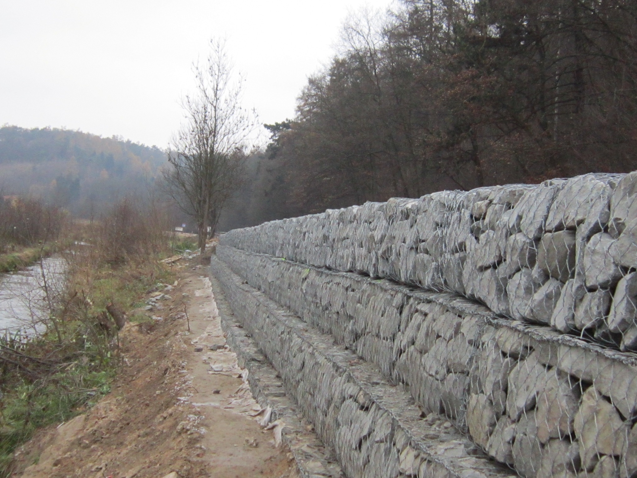 Długa, trójstopniowa ściana oporowa z kamieni w metalowej siatce, biegnąca wzdłuż brzegu rzeki, z lasem w tle i pochmurnym niebem.